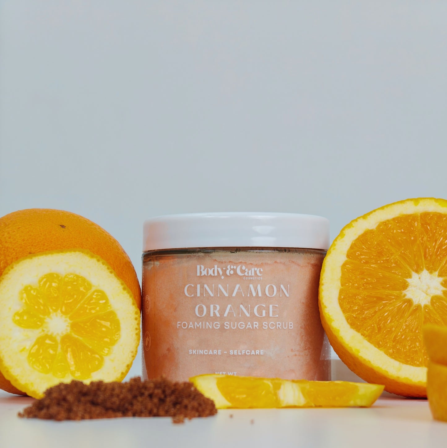 Body & Care Cinnamon Orange Foaming Sugar Scrub in a clear jar, styled with fresh orange halves and orange slices on a white background.