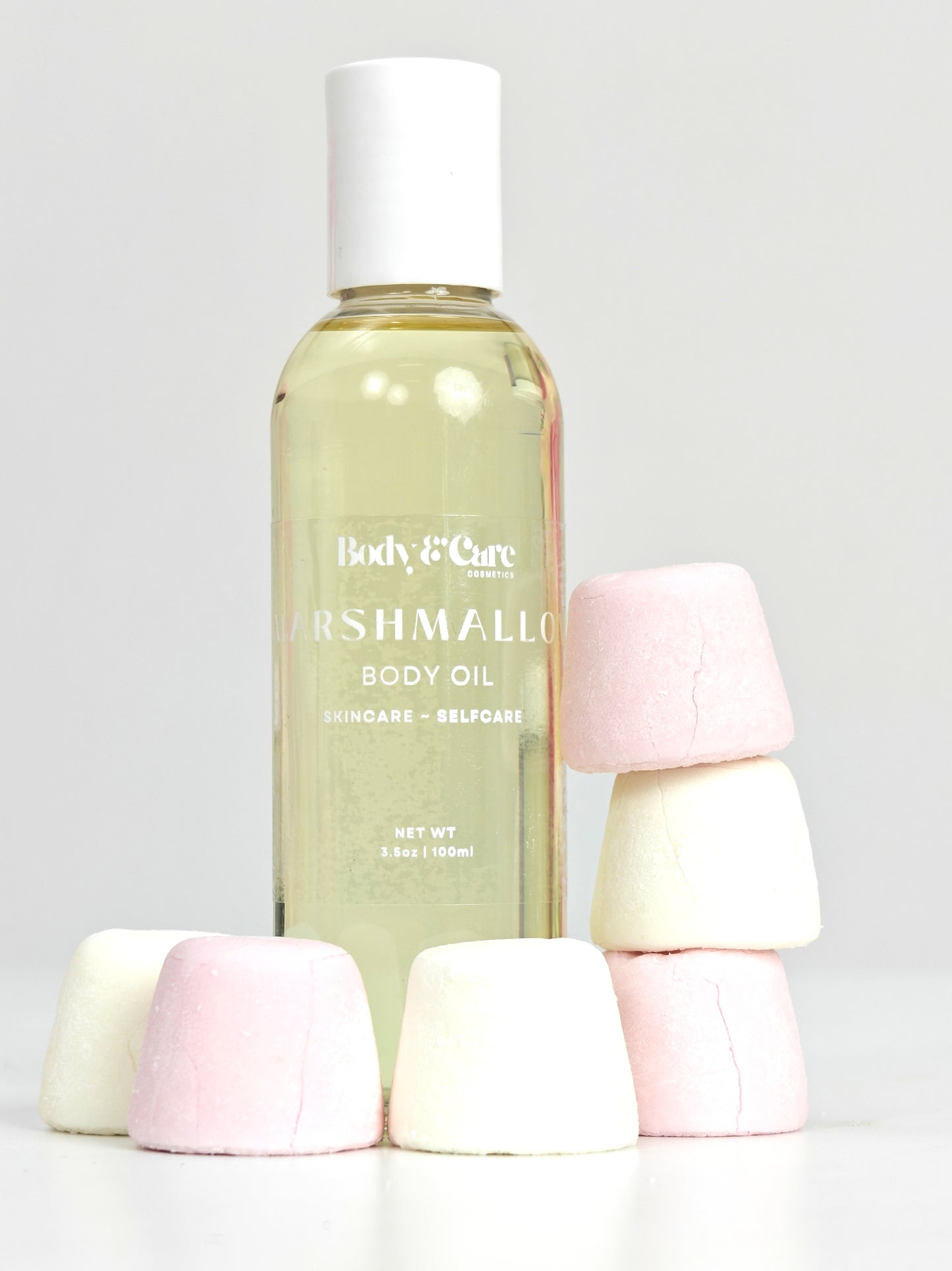 Body & Care Marshmallow Body Oil in a clear bottle with white cap, styled with stacked pink and white marshmallows on a light background.