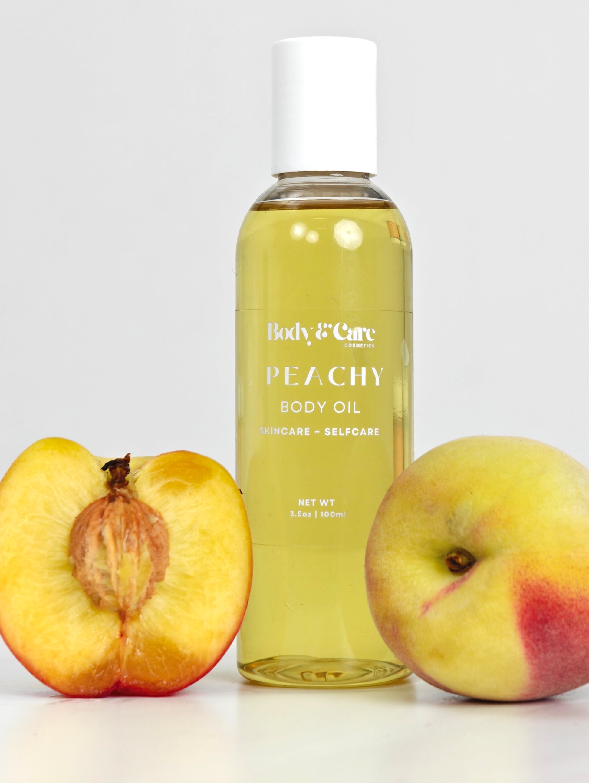Body & Care Peachy Body Oil in a clear bottle with white cap, styled with whole and halved peaches on a light background.