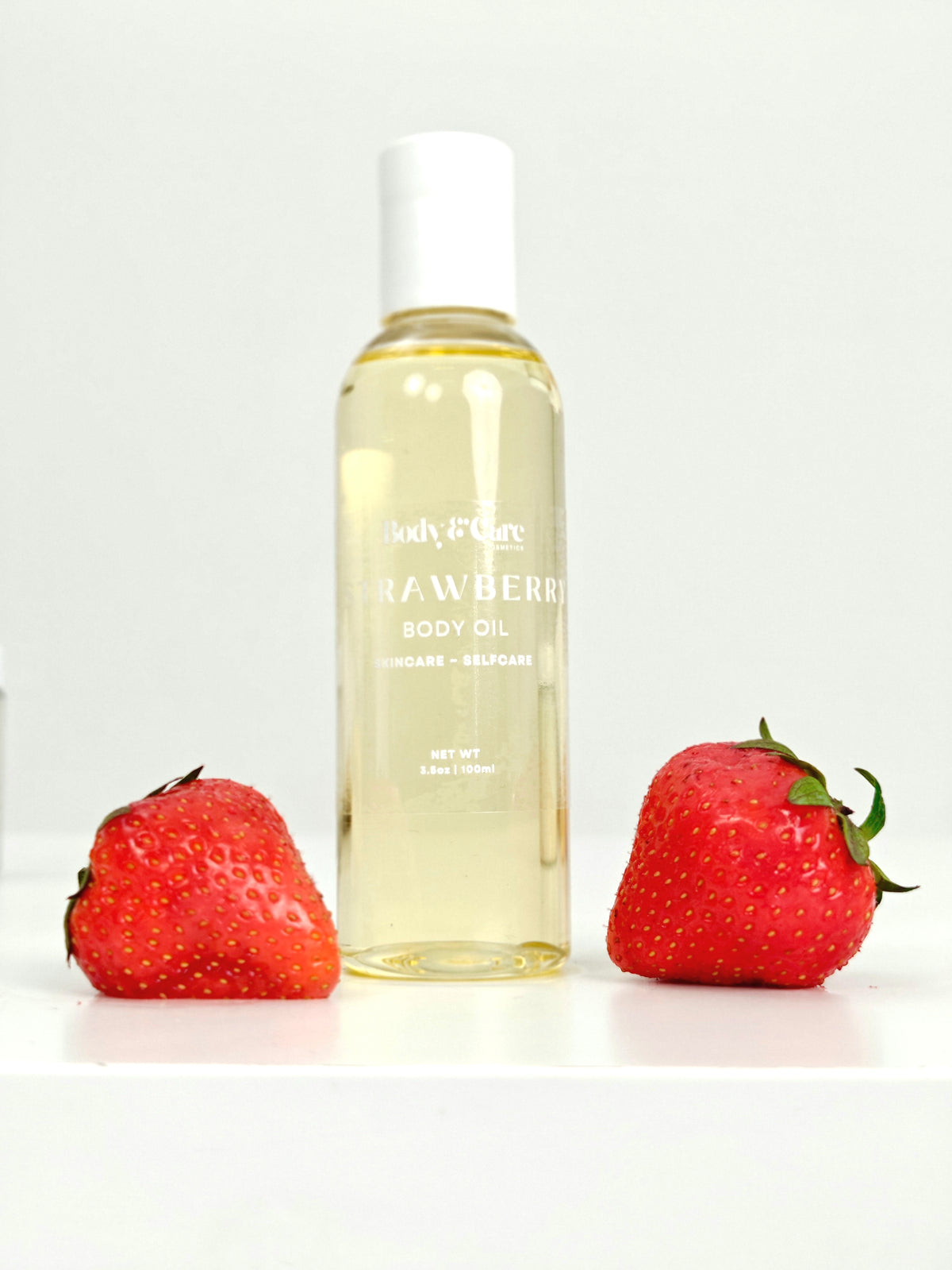 Body & Care Strawberry Body Oil in a clear bottle with white cap, styled with fresh strawberries on a white background.