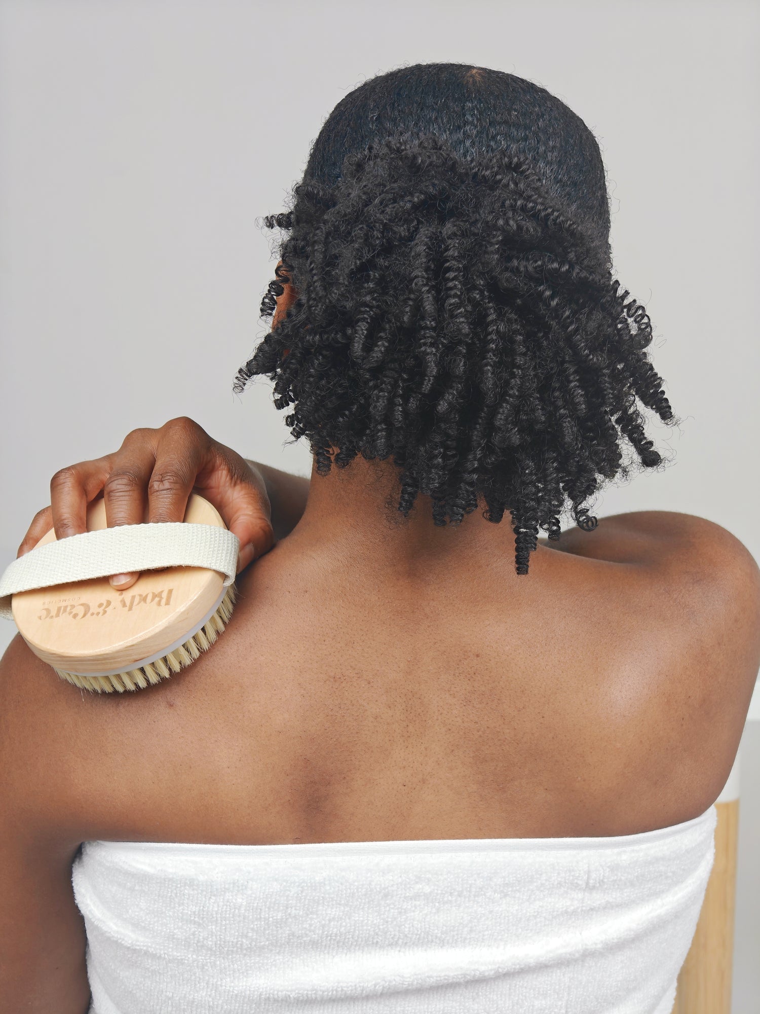 Person using a Body & Care wooden body brush on their back for dry brushing, wrapped in a white towel.