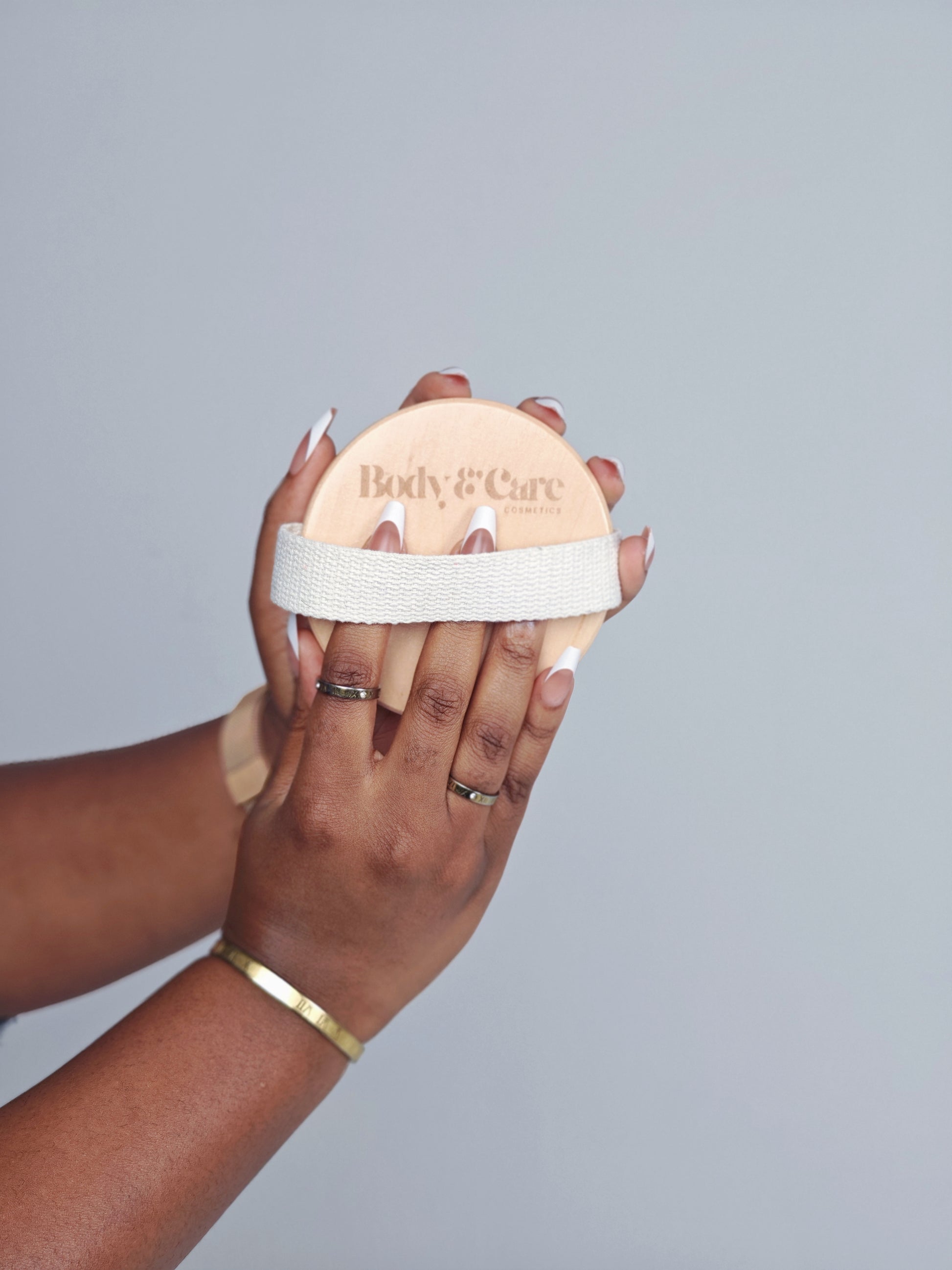 Hands holding a Body & Care wooden body brush with fabric hand strap against a light background.